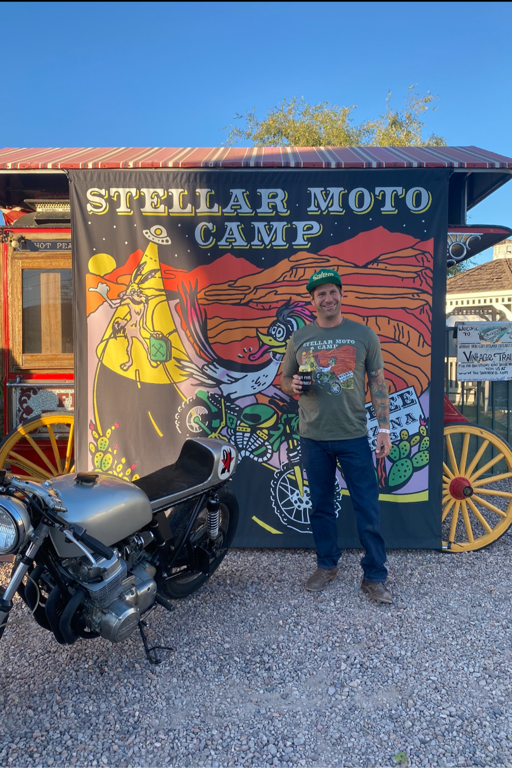 A person standing next to a motorcycle in front of a colorful backdrop with 'STELLAR Moto Camp' text and various vivid designs.
